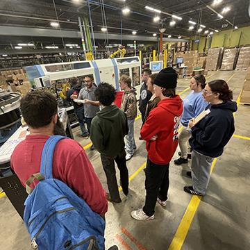 High school students are at a manufacturing facility, facing away from the camera, watching and listening to an employee speaking about his manufacturing job
